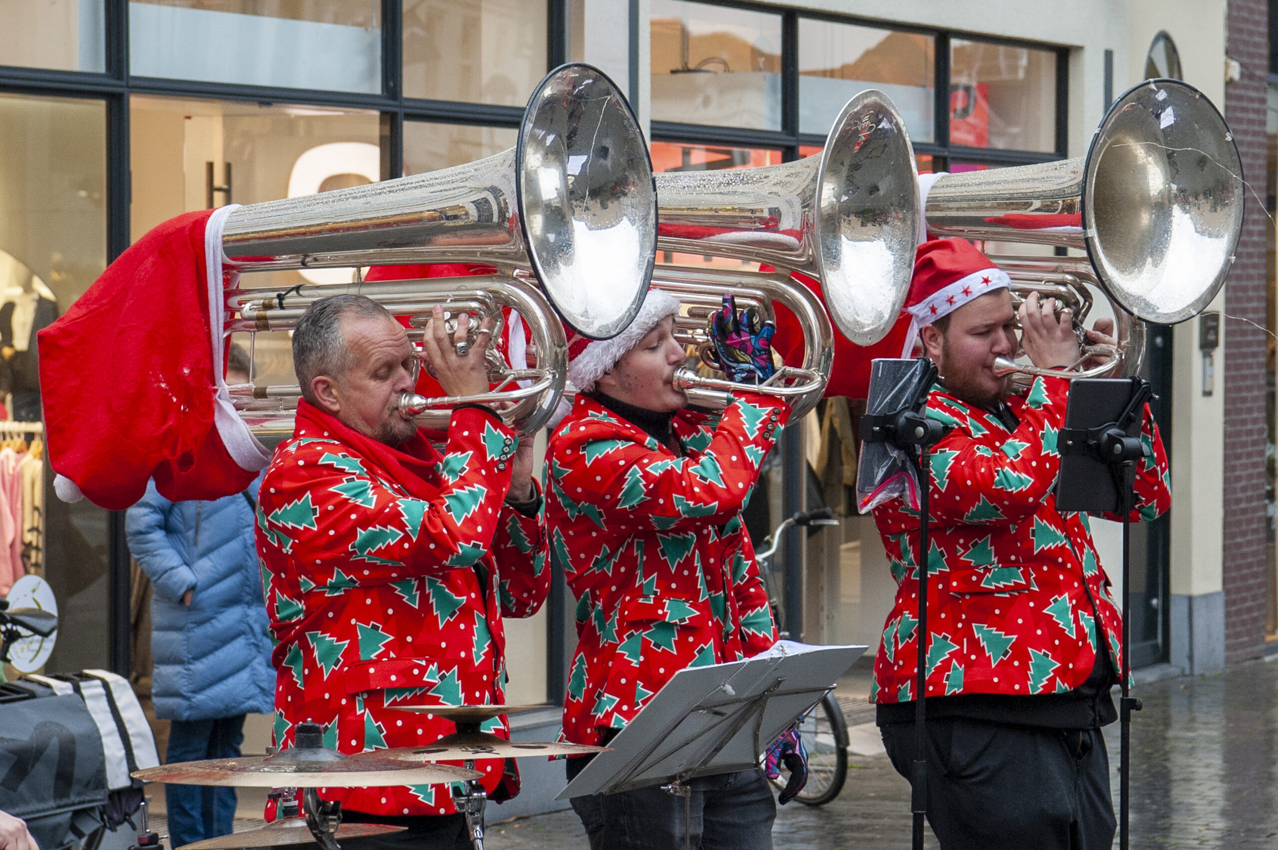 Muzikanten spelen kerstliederen op tubas in een winkelstraat terwijl ze vrolijke kersttruien en kerstmutsen dragen tijdens een feestelijke winterbijeenkomst.