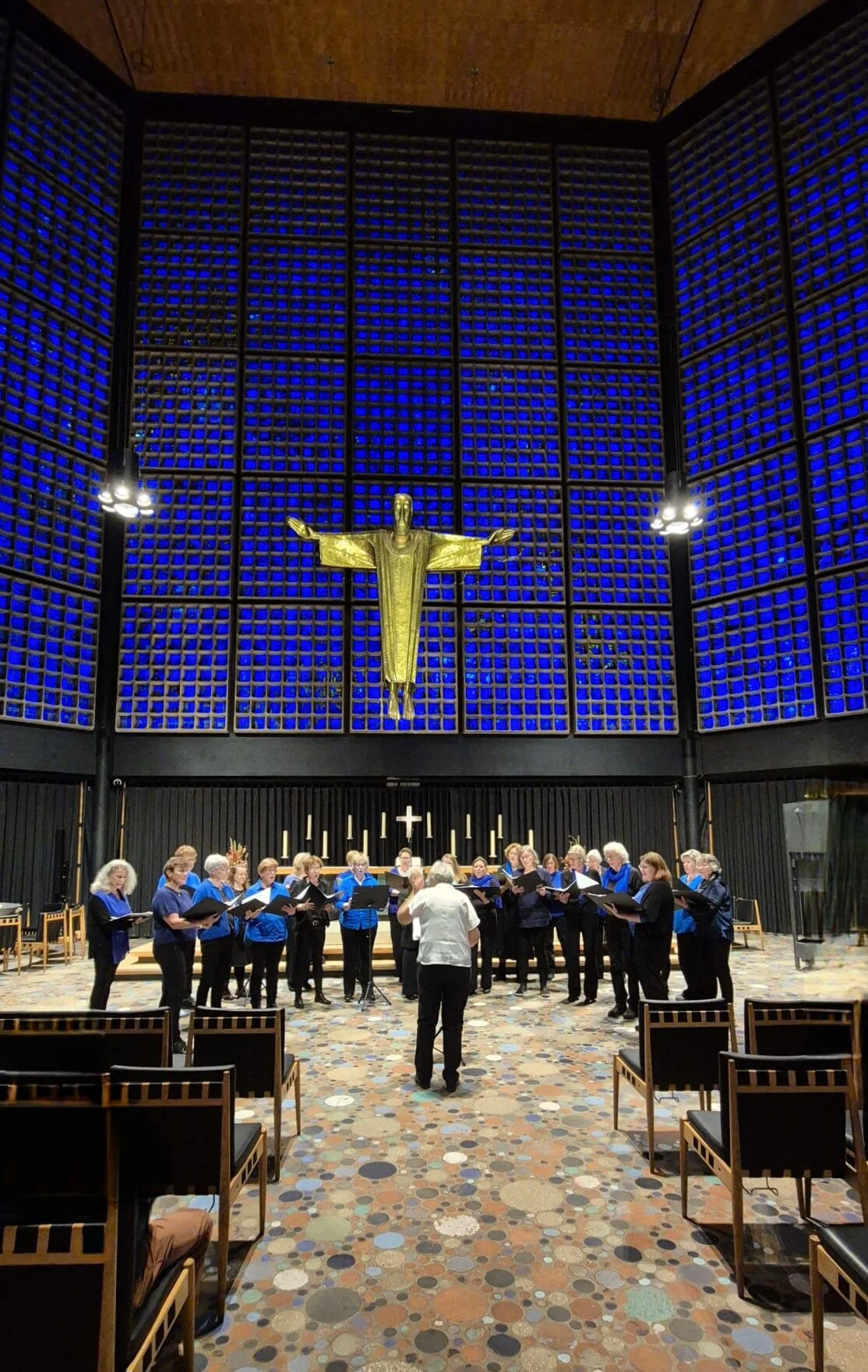 Een koor zingt in een moderne kerk met glazen panelen en een groot goudkleurig kruisbeeld op de achtergrond sfeervolle verlichting en een kleurrijke vloer.