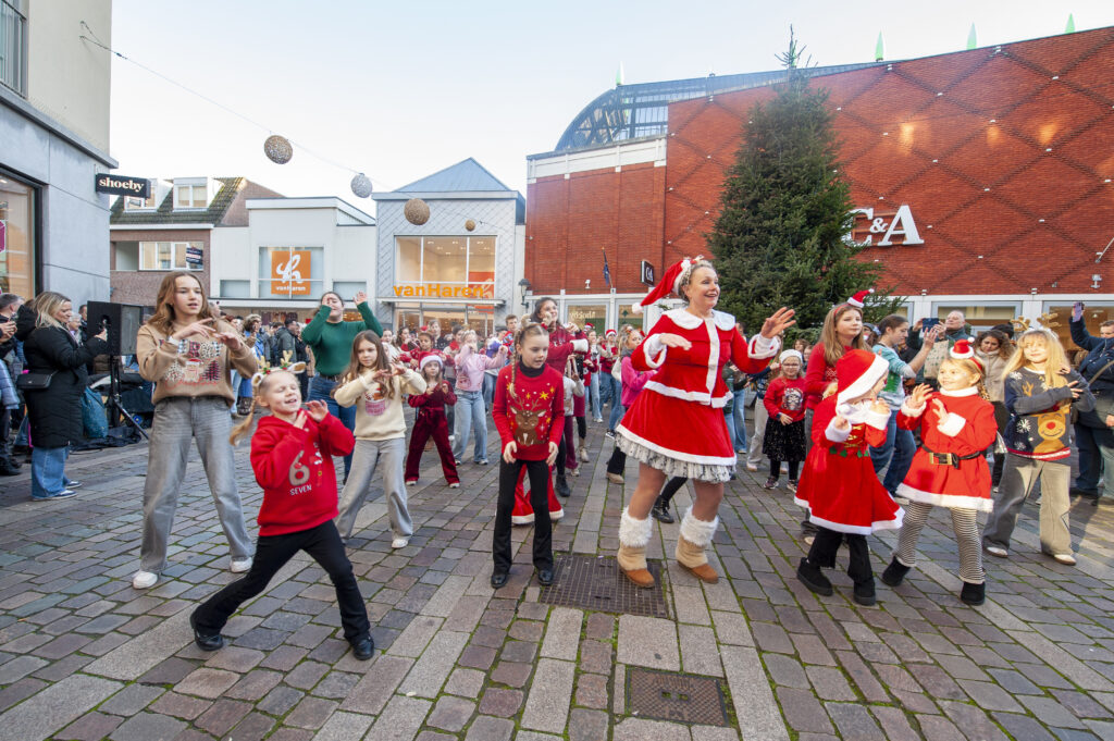 Een groep mensen in feestelijke kerstkleding geniet van een vrolijk samenzijn op een plein in een winkelgebied versierd met een kerstboom en pakjes in de winter.