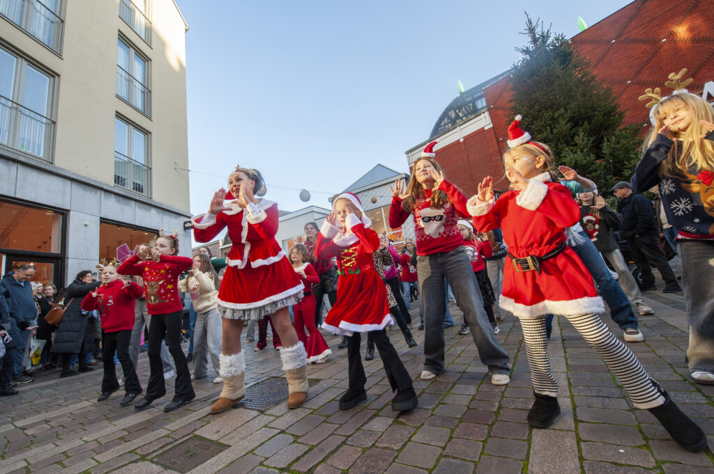 Groep kinderen gekleed in kerstkleding neemt deel aan een vrolijke straatdans tijdens een winterevenement in een stadscentrum met omstanders die toekijken bij zonnig weer.
