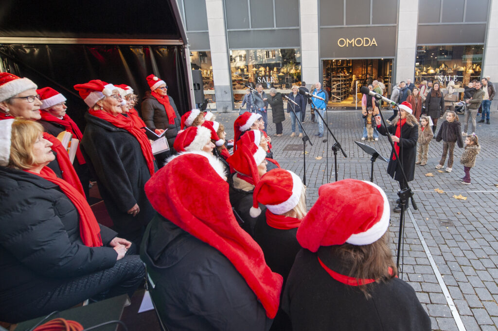Kerstkoor in rode kerstmutsen zingt op een podium in een winkelstraat terwijl voorbijgangers toekijken sfeervolle muzikale viering tijdens de feestdagen in de stad