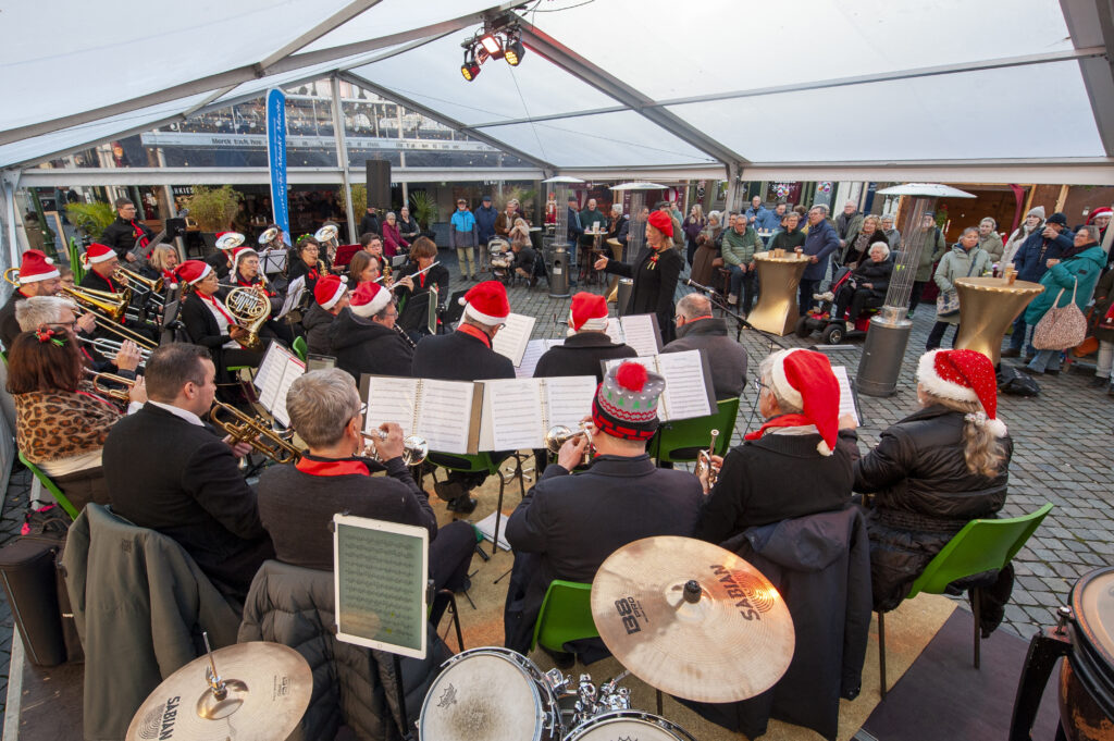 Muzikanten met kerstmutsen spelen in een tent voor een publiek tijdens een kerstconcert op een plein omringd door toeschouwers en feestelijke decoraties.