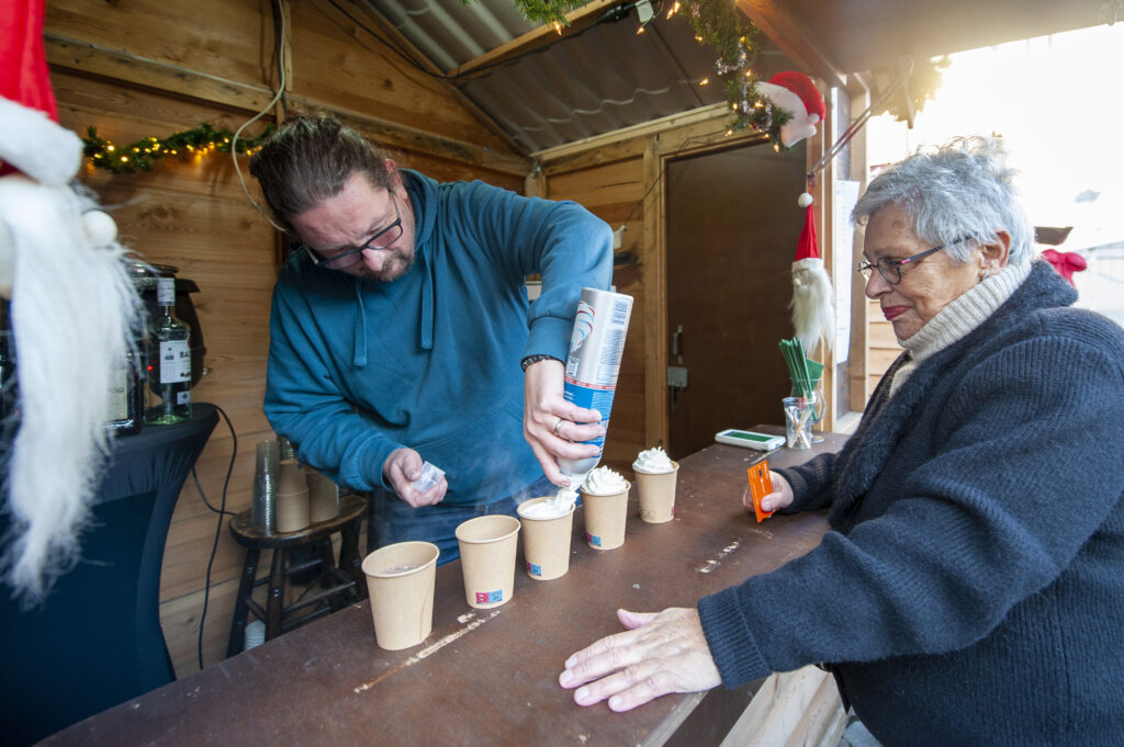 Man schenkt slagroom in bekers op gezellige kerstmarkt terwijl oudere vrouw toekijkt in winterse sfeer met houten kraam en feestelijke decoraties op de achtergrond.