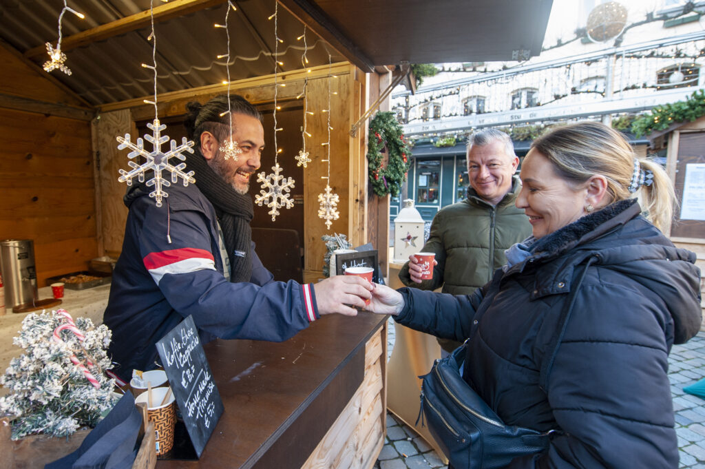 Man bedient klanten in een gezellig houten kerstmarktstalletje versierd met sneeuwvlokken en lichtjes op een koude winterdag terwijl warme drankjes worden geserveerd.