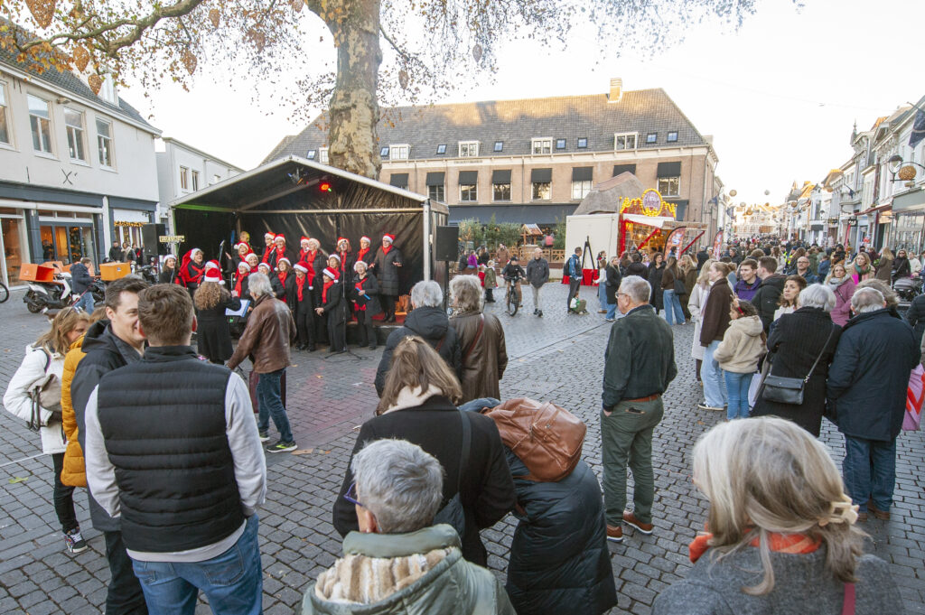 Mensen verzamelen zich op een plein voor een open podium met kerstkoor in rode kleding onder een grote boom tijdens een kerstmarkt in een pittoresk stadscentrum.