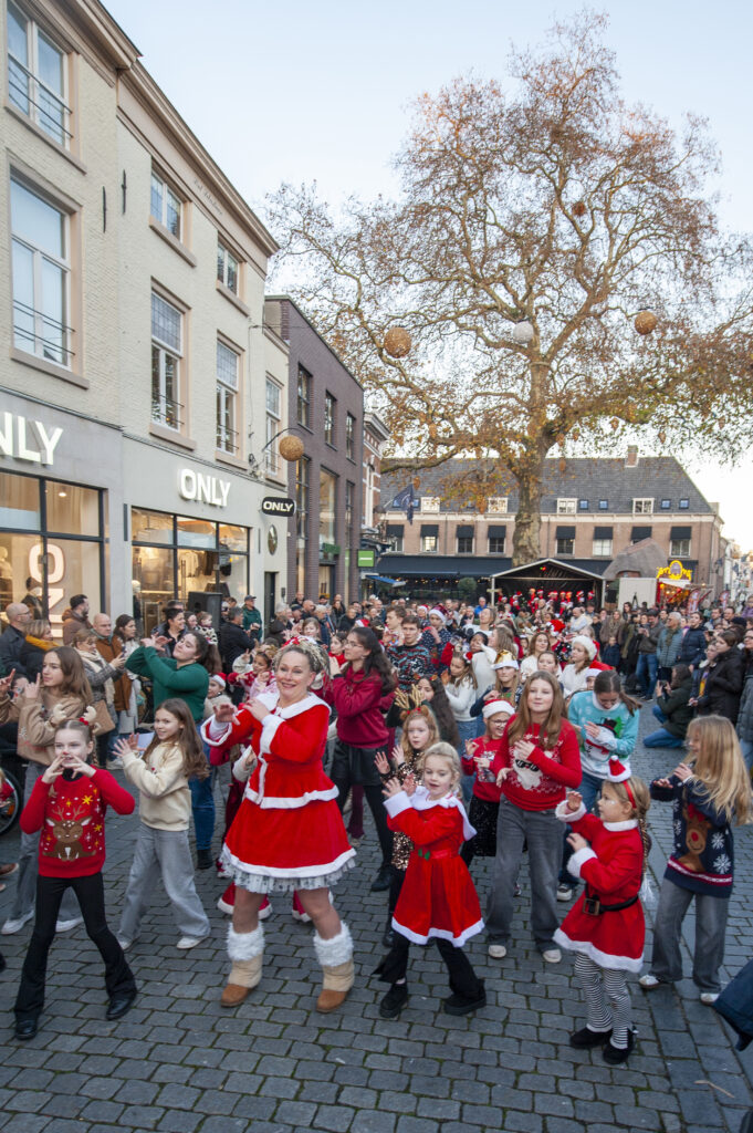 Kerstfeest op straat met mensen in kerstkleding dansend en lachend. Winkels op de achtergrond creëren een feestelijke sfeer in een gezellige winterse setting.
