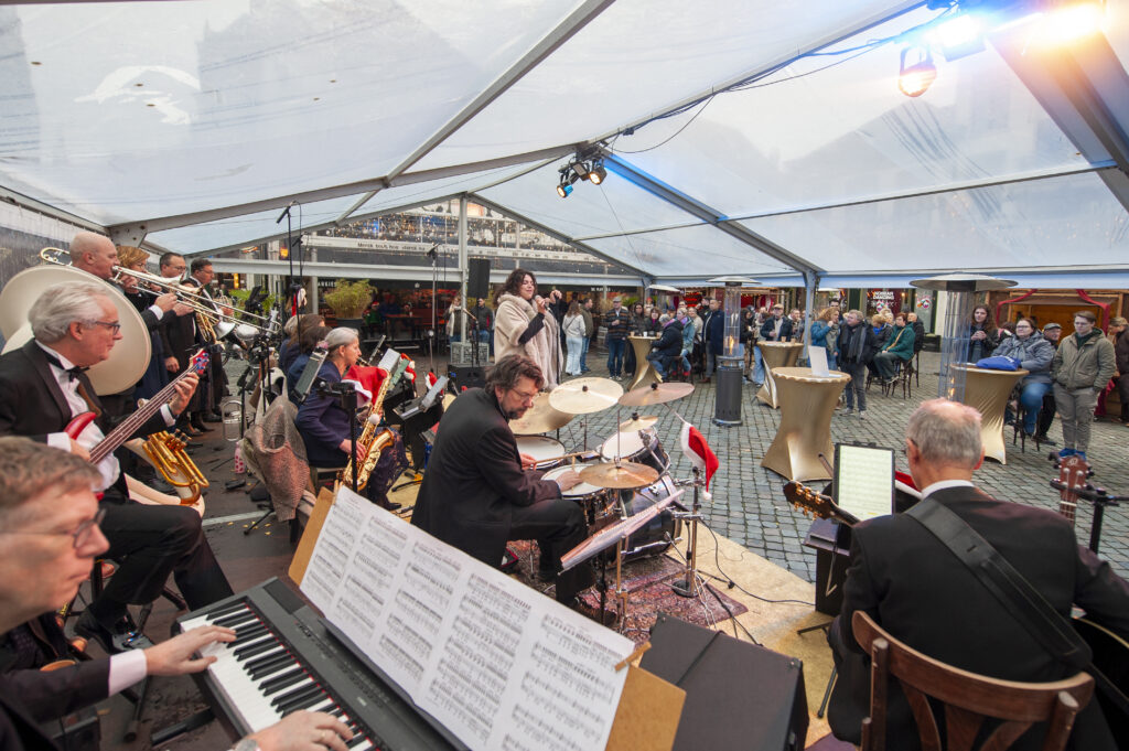 Muzikanten spelen live onder een tent tijdens een evenement met een publiek op de achtergrond. Het orkest zorgt voor een feestelijke sfeer op een plein.
