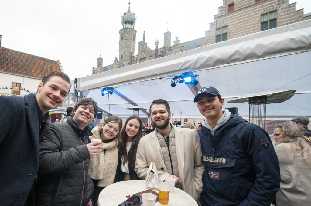 Groep mensen die lachen en poseren voor een camera tijdens een buitenevenement met historische architectuur op de achtergrond en tafels met drankjes.