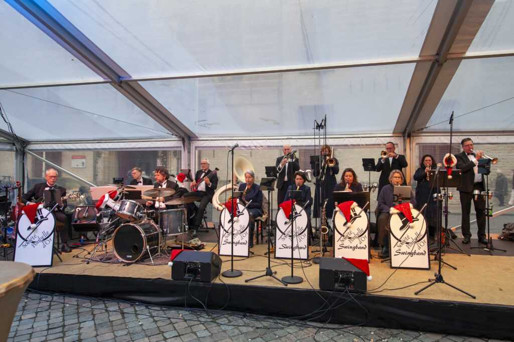 Muziekband speelt live onder een grote overkapping op een podium met diverse instrumenten en muziekstandaards voor een publiek op een geplaveide binnenplaats.