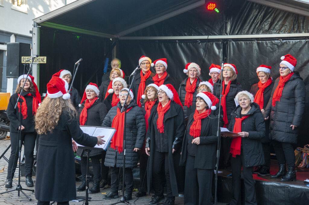 Kerstkoor zingt op een podium tijdens een buitenoptreden. De leden dragen rode sjaals en kerstmutsen. Het is een koude dag met feestelijke sfeer.