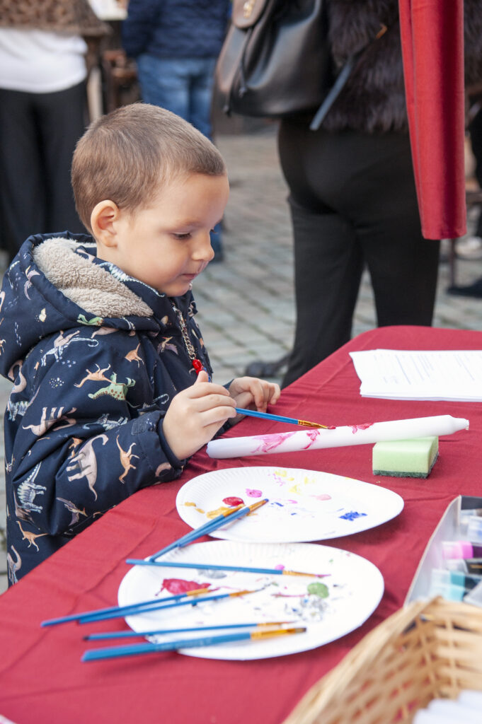 Klein kind schildert met penselen op een kunsttafel in de buitenlucht tijdens een kleinschalig evenement. Tafels bedekt met rood tafelkleed en witte papierborden.
