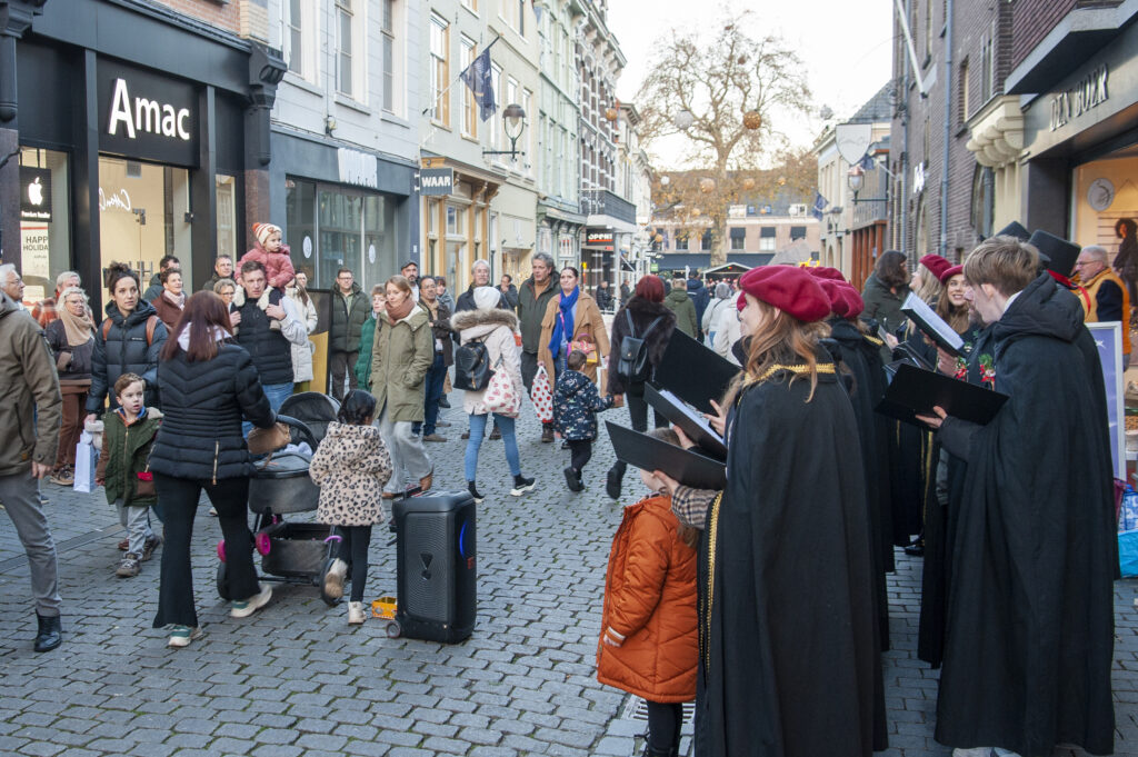 Groep mensen geniet van een koor dat muziek uitvoert op een drukke winkelstraat met passerende voetgangers en winkels op de achtergrond in een sfeervolle stad.