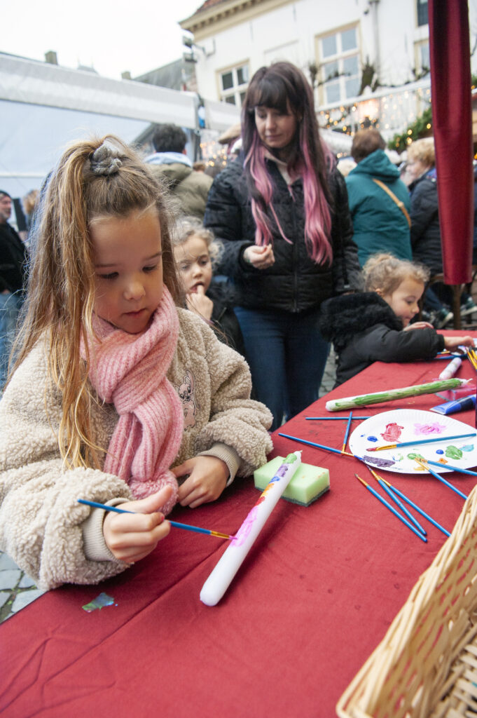 Jong meisje schildert een kaars bij een knutselactiviteit op een wintermarkt met bezoekers in warme kleding op de achtergrond voor een gezellige sfeer in de buitenlucht.
