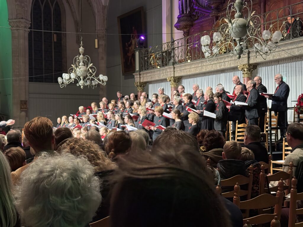 Koor zingt voor publiek in sfeervol verlichte kerk met kroonluchters. Zangers in formele kleding brengen meeslepend optreden in historische omgeving.